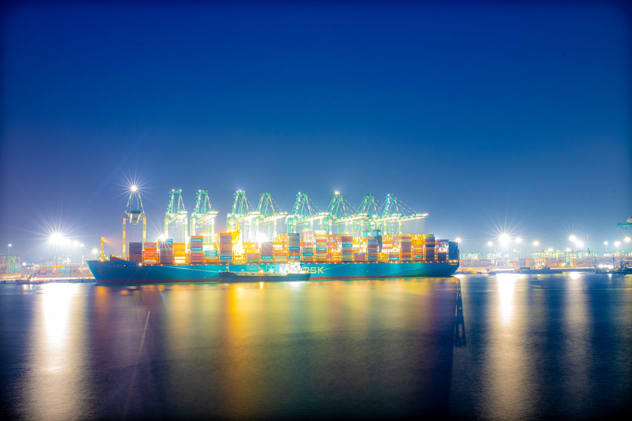 A brightly lit cargo ship with cranes in a bustling harbor at night.